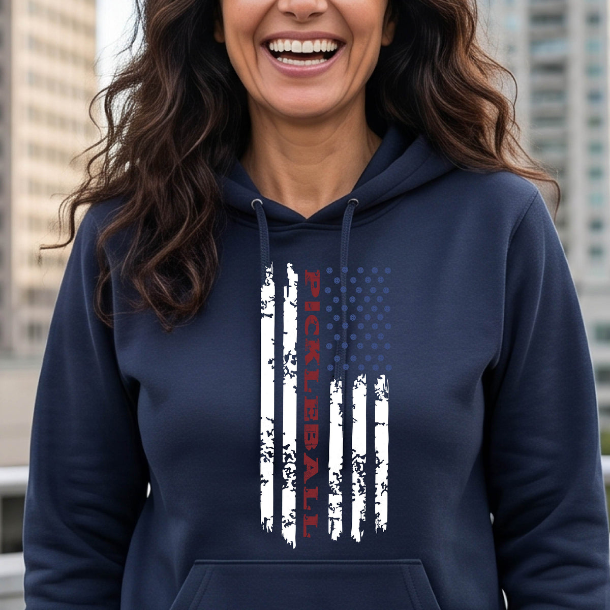 A woman wearing a navy blue fitted hoodie pullover with a pickleball graphic of a distressed flag made out of pickleballs and the text 'PICKLEBALL' replacing one of the stripes, standing on a pickleball court.