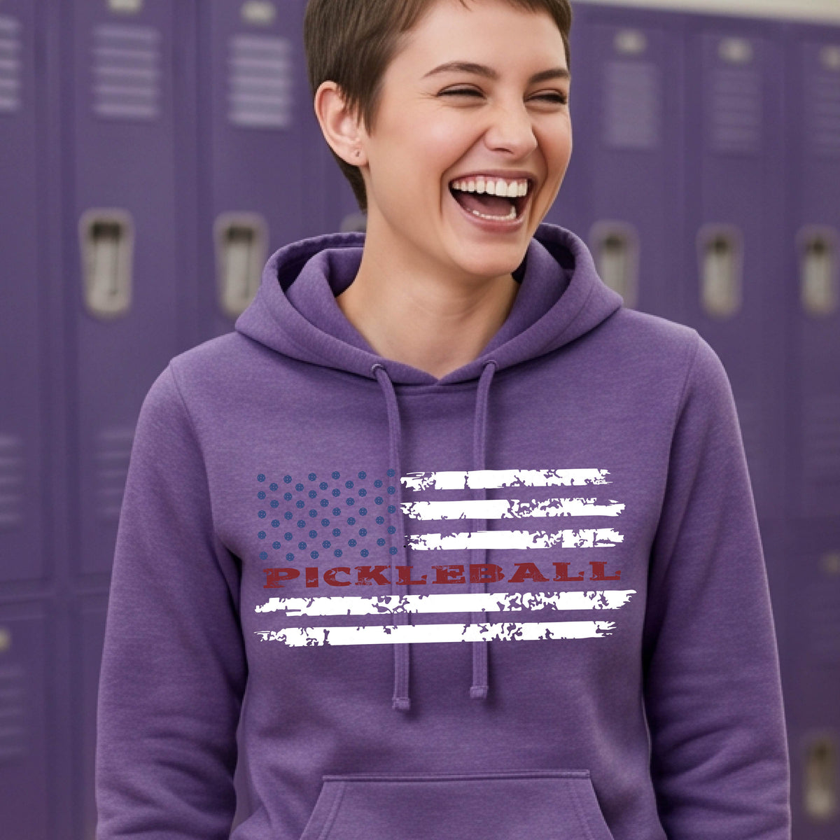 A woman wearing a purple fitted hoodie pullover with a pickleball graphic of a distressed flag made out of pickleballs and the text 'PICKLEBALL' replacing one of the stripes, standing on a pickleball court.