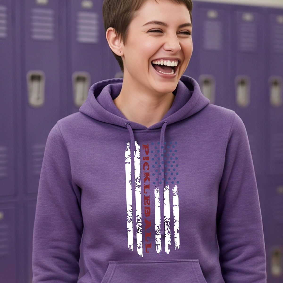 A woman wearing a purple fitted hoodie pullover with a pickleball graphic of a distressed flag made out of pickleballs and the text 'PICKLEBALL' replacing one of the stripes, standing on a pickleball court.