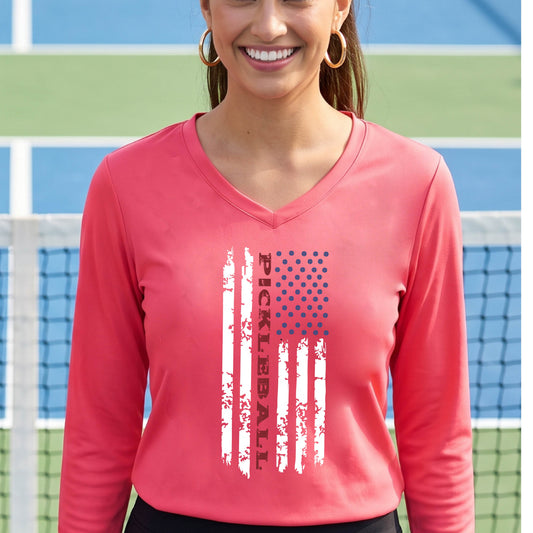 A woman wearing a coral pink long-sleeve t-shirt with a pickleball graphic of a distressed flag made out of pickleballs and the text 'PICKLEBALL' replacing one of the stripes, standing on a pickleball court.