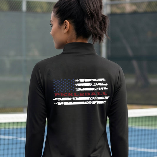 A woman wearing ablack quarter-zip shirt with a pickleball graphic on the back of a distressed flag made out of pickleballs and the text 'PICKLEBALL' replacing one of the stripes, standing on a pickleball court.