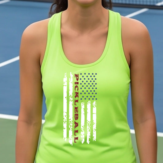 A woman wearing a lime green racerback tank top with a pickleball graphic of a distressed flag made out of pickleballs and the text 'PICKLEBALL' replacing one of the stripes, standing on a pickleball court.