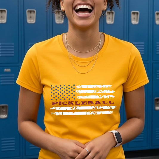 A woman wearing a gold short-sleeve t-shirt with a pickleball graphic of a distressed flag made out of pickleballs and the text 'PICKLEBALL' replacing one of the stripes, standing on a pickleball court.