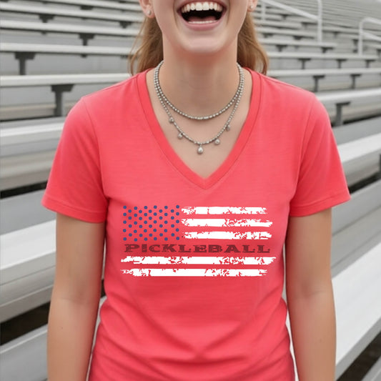 A woman wearing a coral short-sleeve v-neck t-shirt with a pickleball graphic of a distressed flag made out of pickleballs and the text 'PICKLEBALL' replacing one of the stripes, standing on a pickleball court.