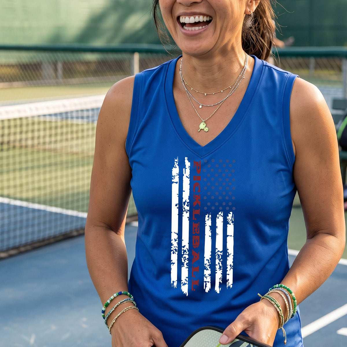 A woman wearing a royal blue sleeveless t-shirt with a pickleball graphic of a distressed flag made out of pickleballs and the text 'PICKLEBALL' replacing one of the stripes, standing on a pickleball court.
