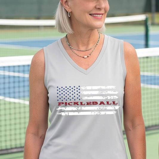 A woman wearing a silver gray sleeveless t-shirt with a pickleball graphic of a distressed flag made out of pickleballs and the text 'PICKLEBALL' replacing one of the stripes, standing on a pickleball court.