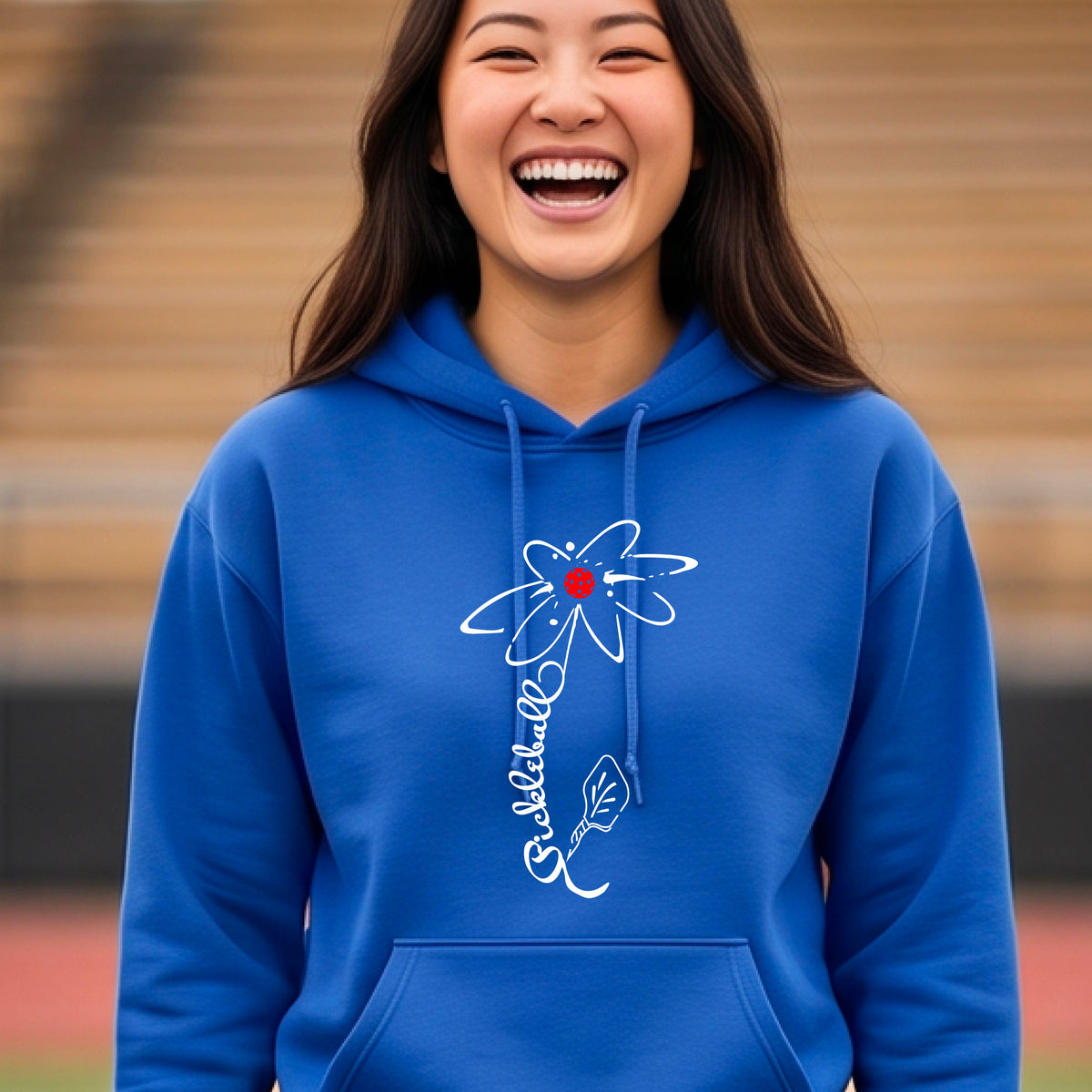 A woman wearing a unisex royal blue hoodie pullover with a pickleball graphic of a flower with a flower that has a colorful pickleball as the center and the stem is made of the word ‘PICKLEBALL’ and the leaf is a pickleball paddle, standing on a pickleball court.
