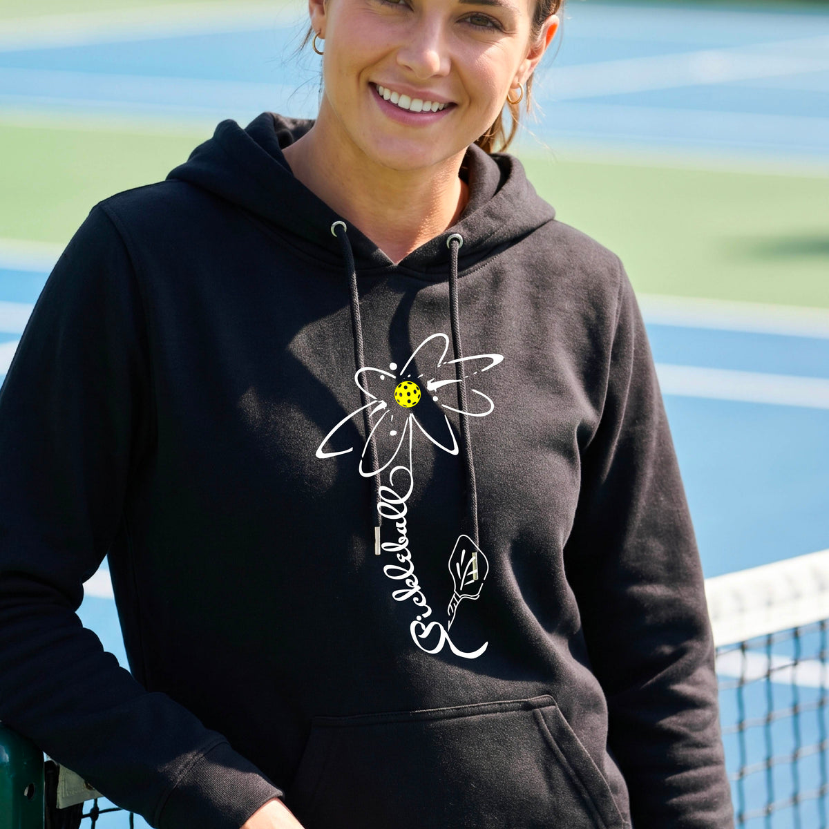 A woman wearing a unisex black hoodie pullover with a pickleball graphic of a flower with a flower that has a colorful pickleball as the center and the stem is made of the word ‘PICKLEBALL’ and the leaf is a pickleball paddle, standing on a pickleball court.