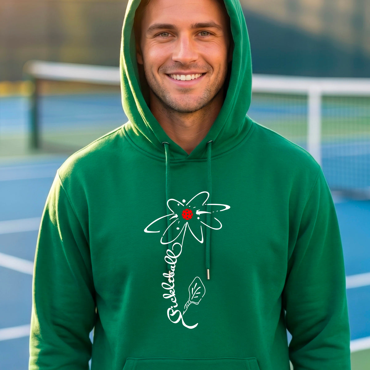 A man wearing a unisex green hoodie pullover with a pickleball graphic of a flower with a flower that has a colorful pickleball as the center and the stem is made of the word ‘PICKLEBALL’ and the leaf is a pickleball paddle, standing on a pickleball court.