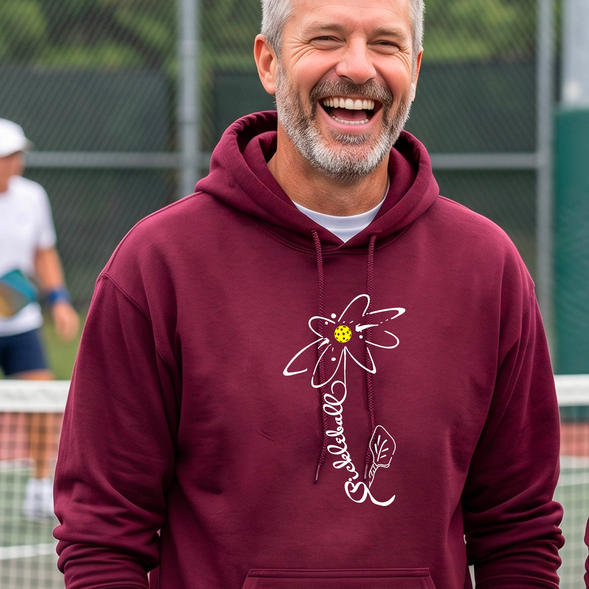 A man wearing a unisex maroon hoodie pullover with a pickleball graphic of a flower with a flower that has a colorful pickleball as the center and the stem is made of the word ‘PICKLEBALL’ and the leaf is a pickleball paddle, standing on a pickleball court.