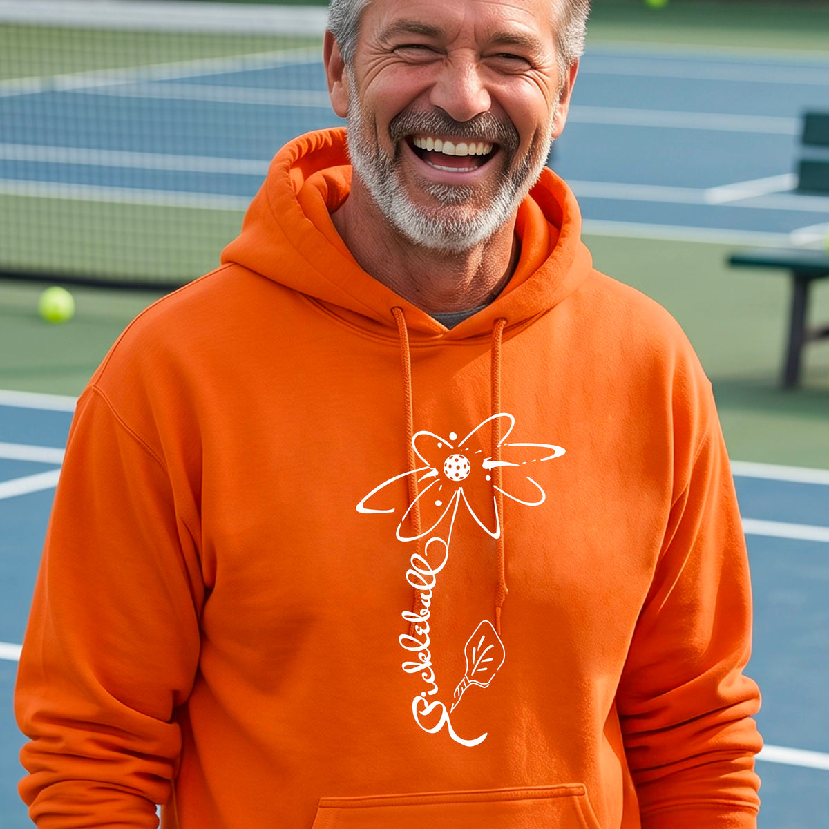 A man wearing a unisex orange hoodie pullover with a pickleball graphic of a flower with a flower that has a colorful pickleball as the center and the stem is made of the word ‘PICKLEBALL’ and the leaf is a pickleball paddle, standing on a pickleball court.