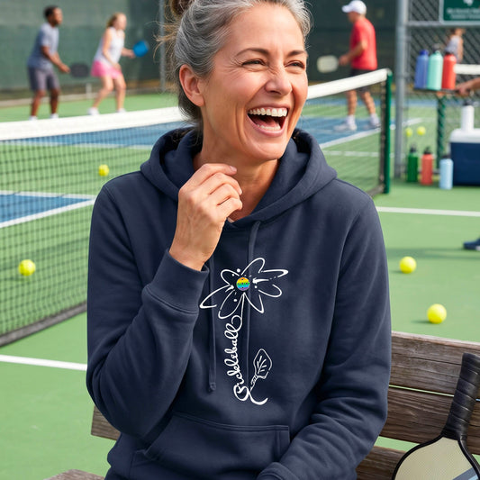 A woman wearing navy blue fitted hoodie pullover with a pickleball graphic of a flower with a flower that has a colorful pickleball as the center and the stem is made of the word ‘PICKLEBALL’ and the leaf is a pickleball paddle, standing on a pickleball court.