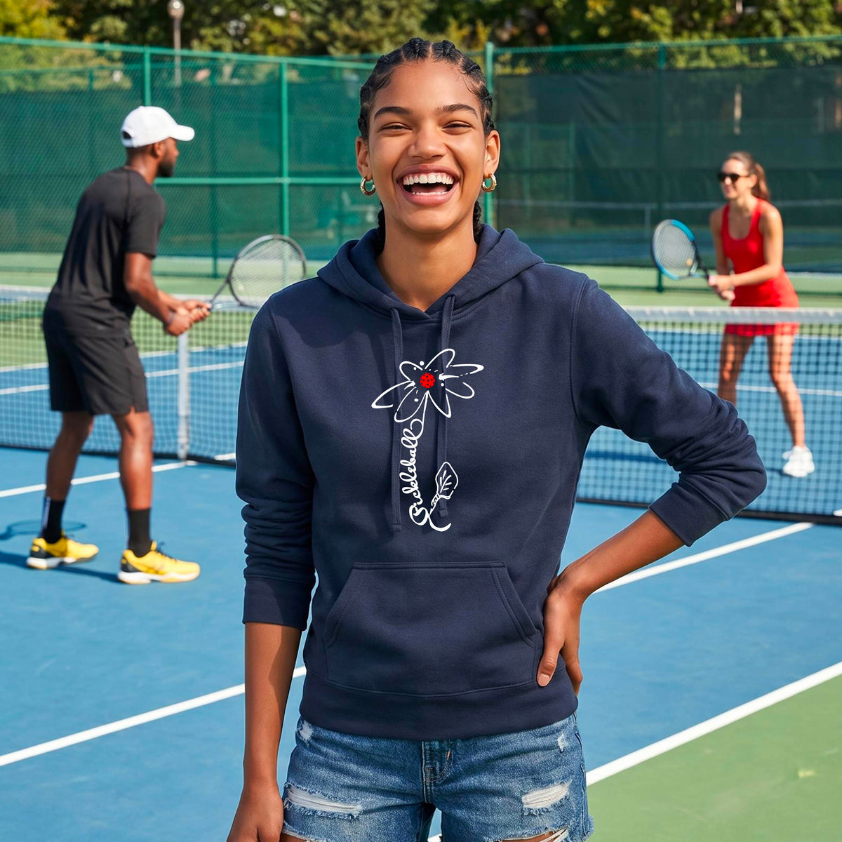 A woman wearing a navy blue fitted hoodie pullover with a pickleball graphic of a flower with a flower that has a colorful pickleball as the center and the stem is made of the word ‘PICKLEBALL’ and the leaf is a pickleball paddle, standing on a pickleball court.