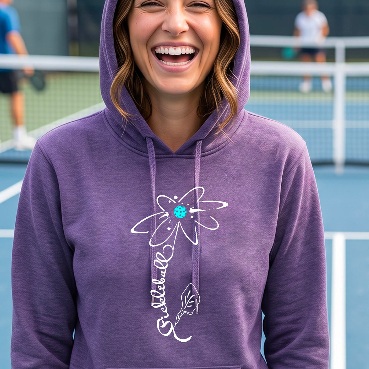 A woman wearing purple fitted hoodie pullover with a pickleball graphic of a flower with a flower that has a colorful pickleball as the center and the stem is made of the word ‘PICKLEBALL’ and the leaf is a pickleball paddle, standing on a pickleball court.