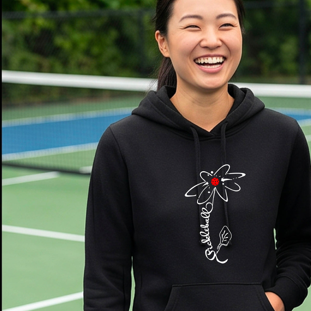 A woman wearing black fitted hoodie pullover with a pickleball graphic of a flower with a flower that has a colorful pickleball as the center and the stem is made of the word ‘PICKLEBALL’ and the leaf is a pickleball paddle, standing on a pickleball court.
