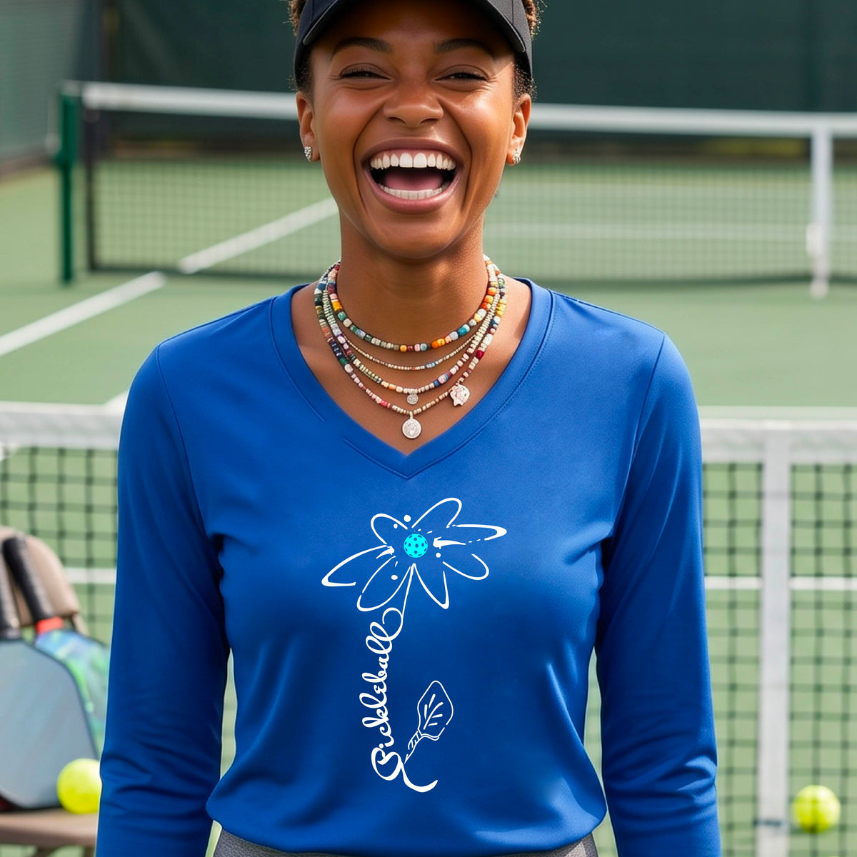A woman wearing a royal blue long-sleeve t-shirt with a pickleball graphic of a flower with a flower that has a colorful pickleball as the center and the stem is made of the word ‘PICKLEBALL’ and the leaf is a pickleball paddle, standing on a pickleball court.