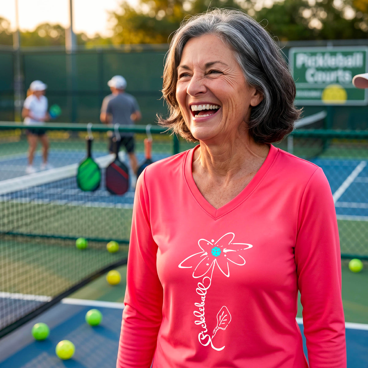 A woman wearing a coral pink long-sleeve t-shirt with a pickleball graphic of a flower with a flower that has a colorful pickleball as the center and the stem is made of the word ‘PICKLEBALL’ and the leaf is a pickleball paddle, standing on a pickleball court.