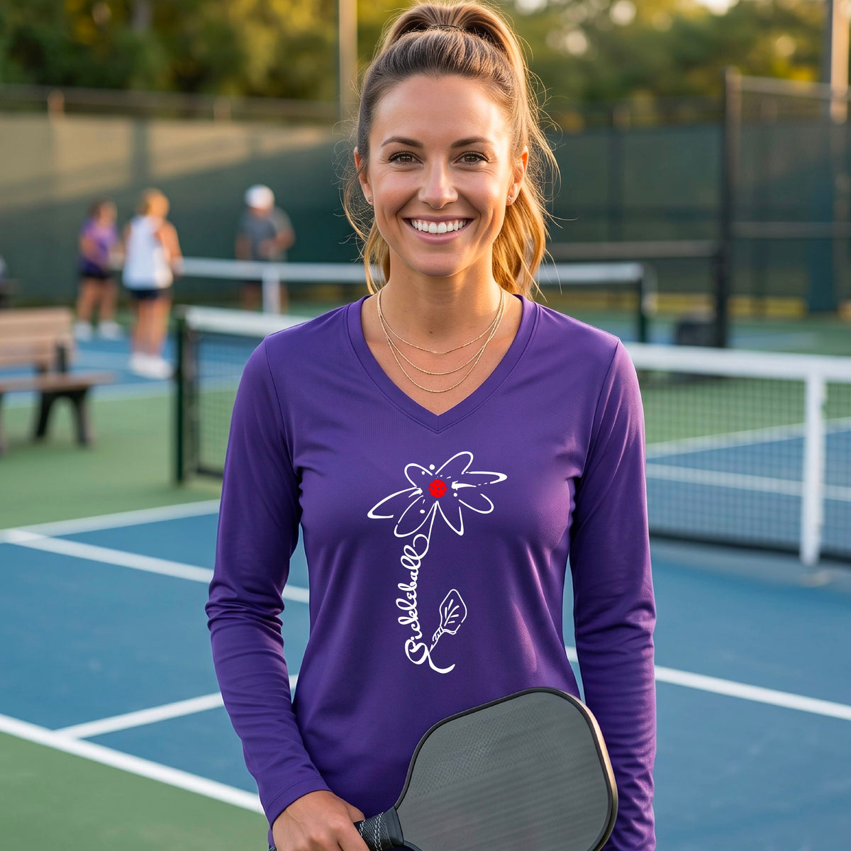 A woman wearing a purple long-sleeve t-shirt with a pickleball graphic of a flower with a flower that has a colorful pickleball as the center and the stem is made of the word ‘PICKLEBALL’ and the leaf is a pickleball paddle, standing on a pickleball court.