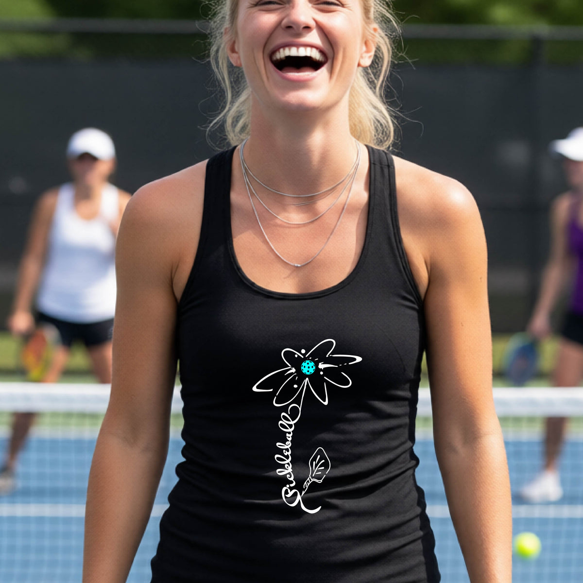 A woman wearing a black racerback tank top with a pickleball graphic of a flower with a flower that has a colorful pickleball as the center and the stem is made of the word ‘PICKLEBALL’ and the leaf is a pickleball paddle, standing on a pickleball court.