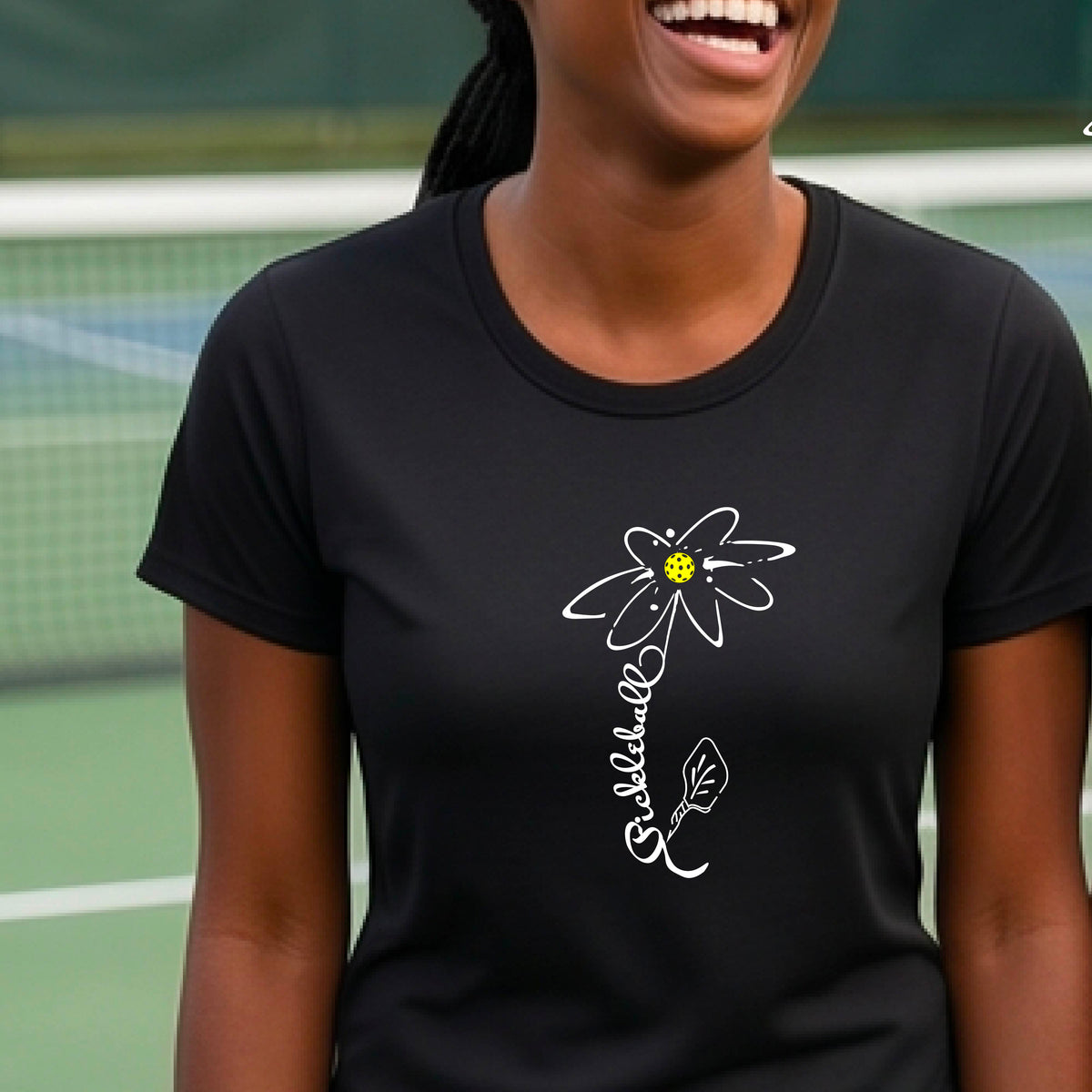 A woman wearing a black short-sleeve t-shirt with a pickleball graphic of a flower with a flower that has a colorful pickleball as the center and the stem is made of the word ‘PICKLEBALL’ and the leaf is a pickleball paddle, standing on a pickleball court.