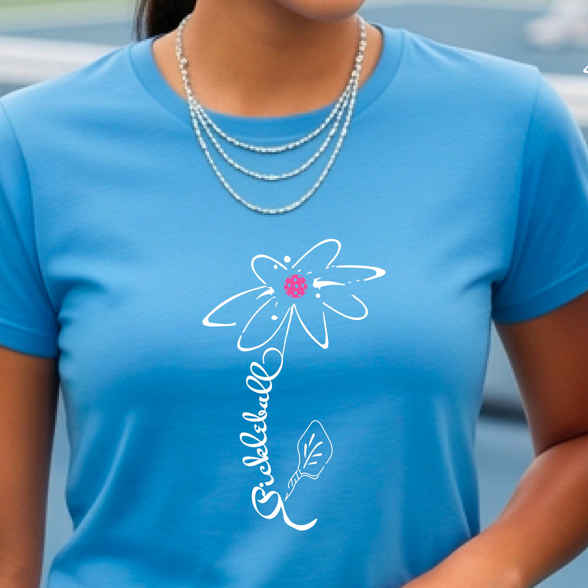 A woman wearing a blue short-sleeve t-shirt with a pickleball graphic of a flower with a flower that has a colorful pickleball as the center and the stem is made of the word ‘PICKLEBALL’ and the leaf is a pickleball paddle, standing on a pickleball court.