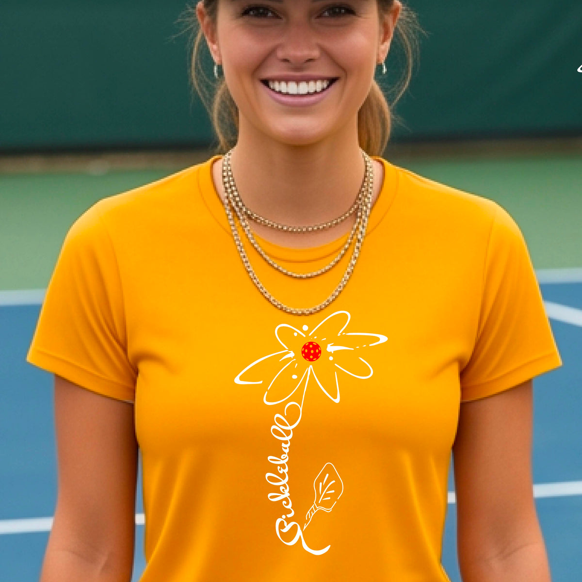 A woman wearing a gold short-sleeve t-shirt with a pickleball graphic of a flower with a flower that has a colorful pickleball as the center and the stem is made of the word ‘PICKLEBALL’ and the leaf is a pickleball paddle, standing on a pickleball court.