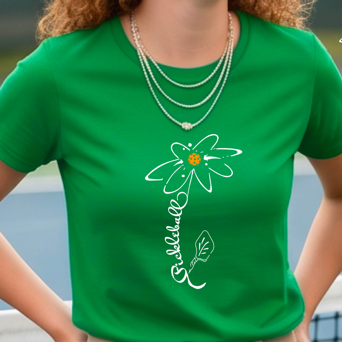 A woman wearing a green short-sleeve t-shirt with a pickleball graphic of a flower with a flower that has a colorful pickleball as the center and the stem is made of the word ‘PICKLEBALL’ and the leaf is a pickleball paddle, standing on a pickleball court.