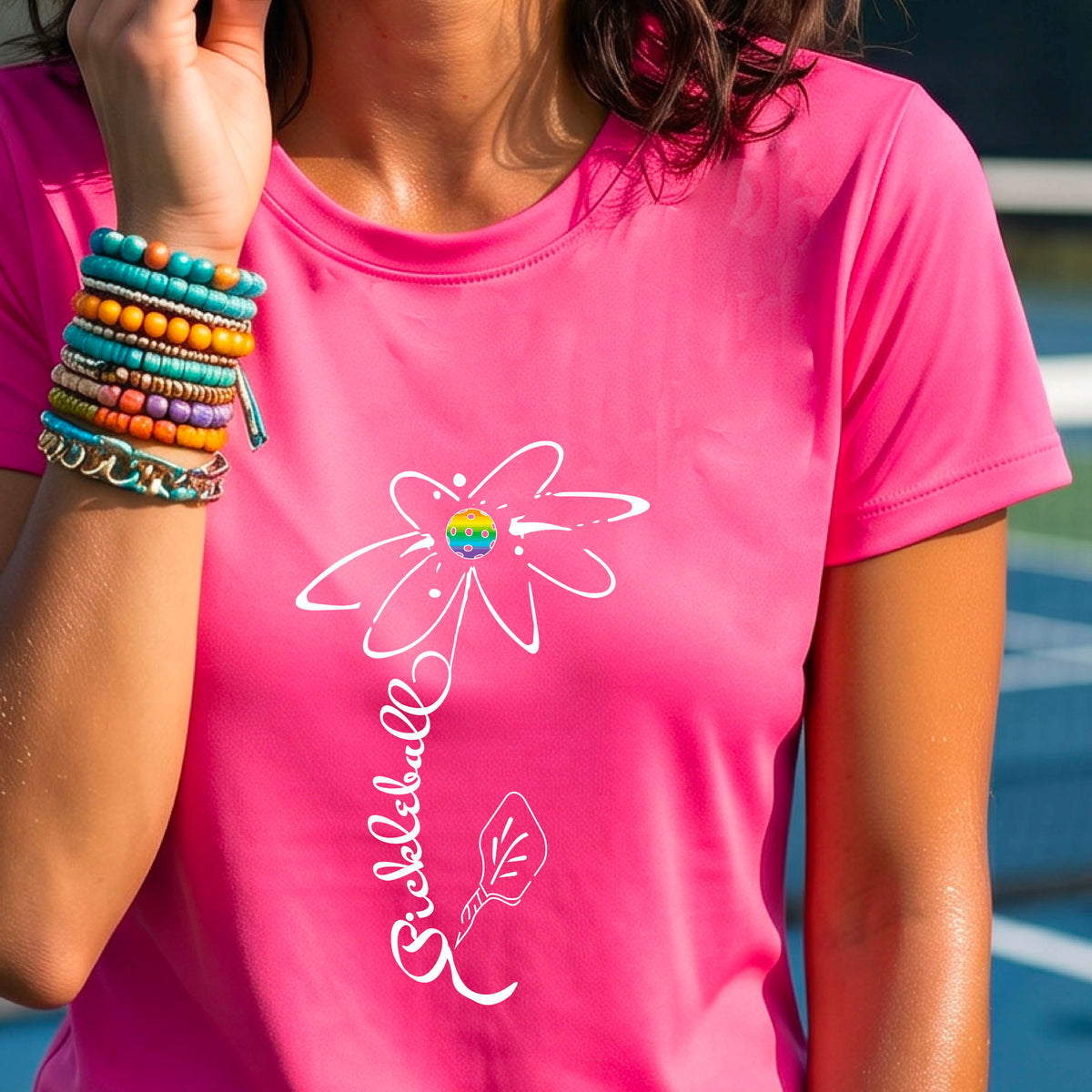 A woman wearing a pink short-sleeve t-shirt with a pickleball graphic of a flower with a flower that has a colorful pickleball as the center and the stem is made of the word ‘PICKLEBALL’ and the leaf is a pickleball paddle, standing on a pickleball court.