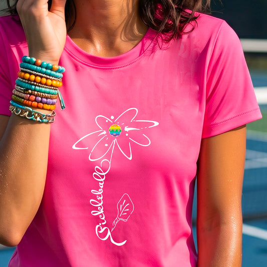 A woman wearing a pink short-sleeve t-shirt with a pickleball graphic of a flower with a flower that has a colorful pickleball as the center and the stem is made of the word ‘PICKLEBALL’ and the leaf is a pickleball paddle, standing on a pickleball court.