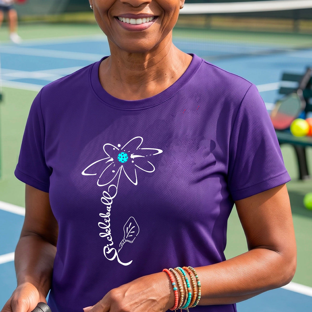 A woman wearing a purple short-sleeve t-shirt with a pickleball graphic of a flower with a flower that has a colorful pickleball as the center and the stem is made of the word ‘PICKLEBALL’ and the leaf is a pickleball paddle, standing on a pickleball court.