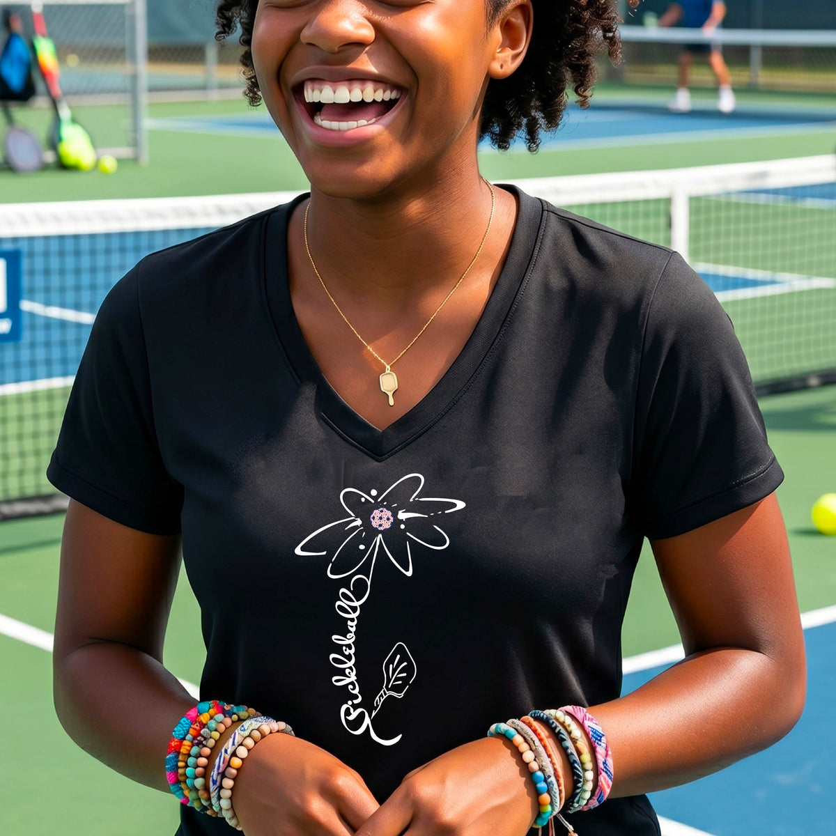 A woman wearing a black short-sleeve V-Neck t-shirt with a pickleball graphic of a flower with a flower that has a colorful pickleball as the center and the stem is made of the word ‘PICKLEBALL’ and the leaf is a pickleball paddle, standing on a pickleball court.