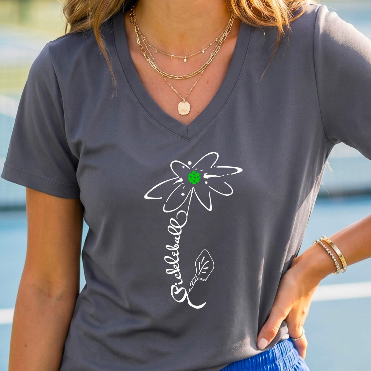 A woman wearing a dark gray short-sleeve V-Neck t-shirt with a pickleball graphic of a flower with a flower that has a colorful pickleball as the center and the stem is made of the word ‘PICKLEBALL’ and the leaf is a pickleball paddle, standing on a pickleball court.