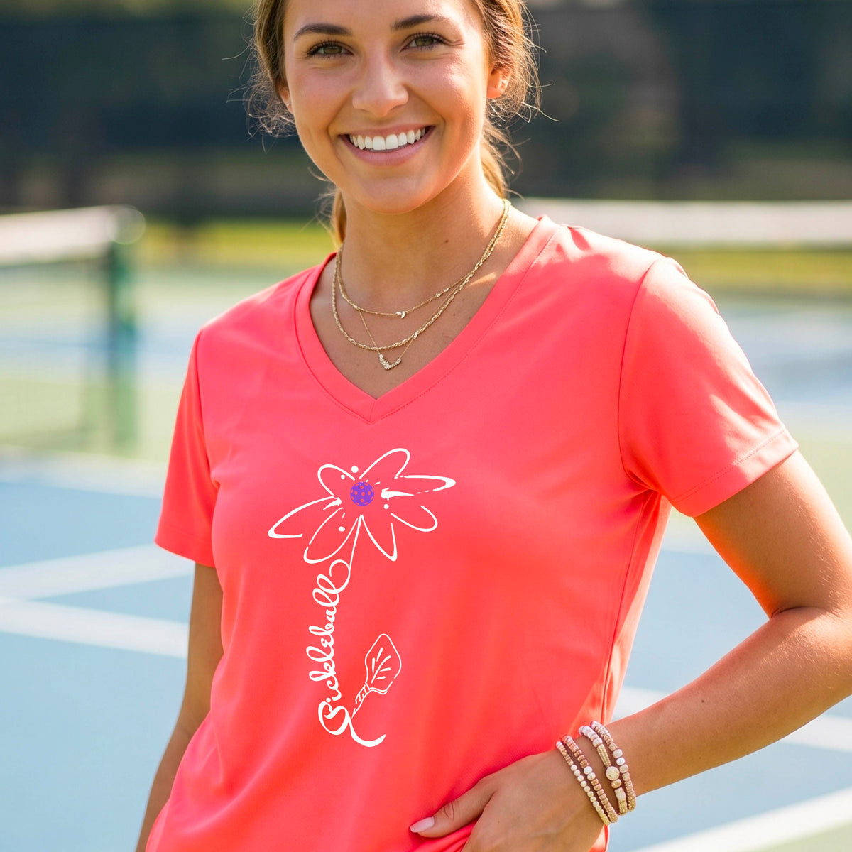 A woman wearing a coral pink short-sleeve V-Neck t-shirt with a pickleball graphic of a flower with a flower that has a colorful pickleball as the center and the stem is made of the word ‘PICKLEBALL’ and the leaf is a pickleball paddle, standing on a pickleball court.