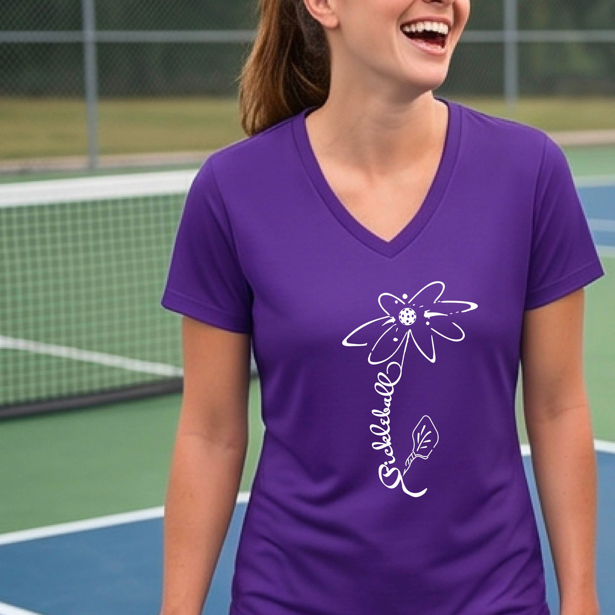 A woman wearing a purple short-sleeve V-Neck t-shirt with a pickleball graphic of a flower with a flower that has a colorful pickleball as the center and the stem is made of the word ‘PICKLEBALL’ and the leaf is a pickleball paddle, standing on a pickleball court.