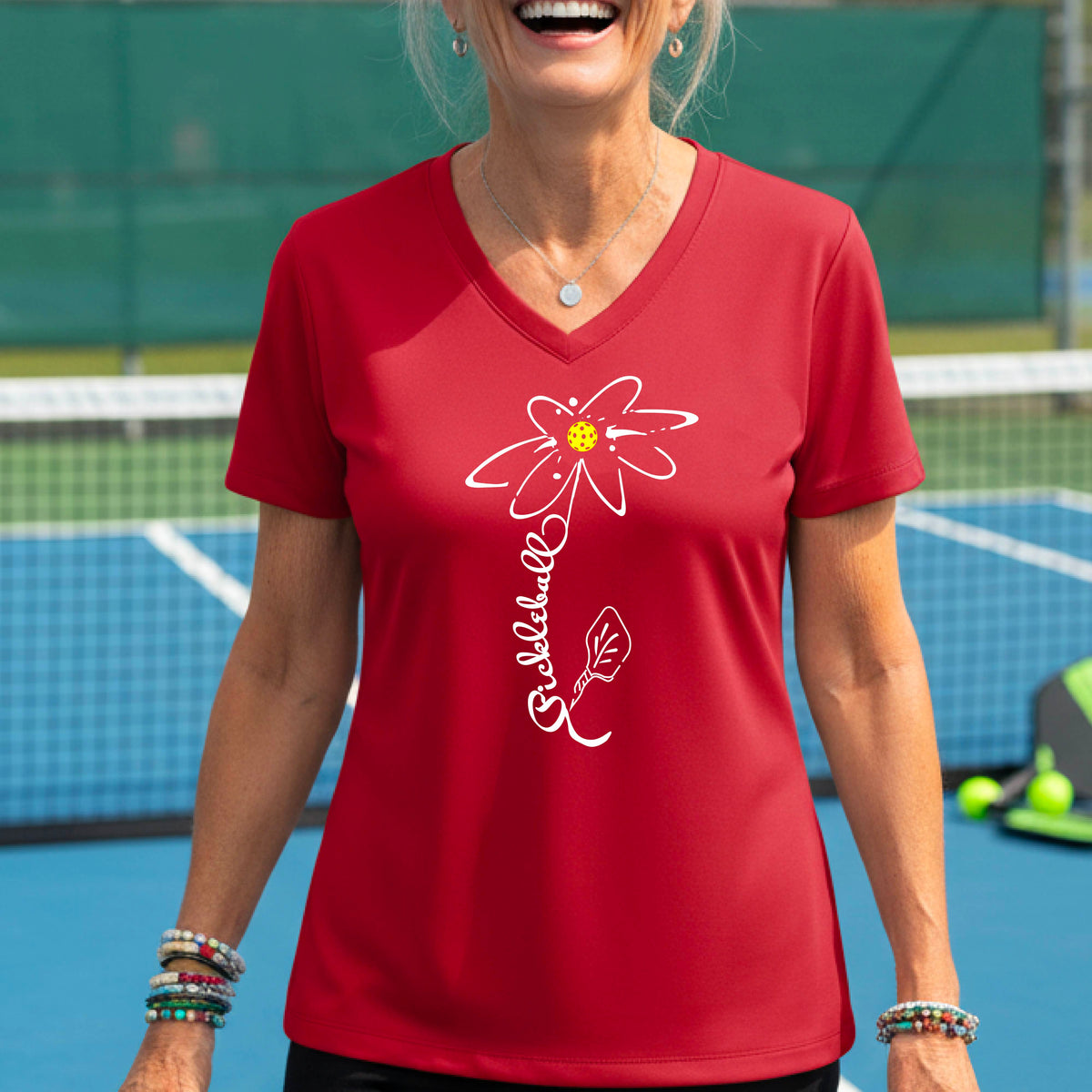 A woman wearing a red short-sleeve V-Neck t-shirt with a pickleball graphic of a flower with a flower that has a colorful pickleball as the center and the stem is made of the word ‘PICKLEBALL’ and the leaf is a pickleball paddle, standing on a pickleball court.