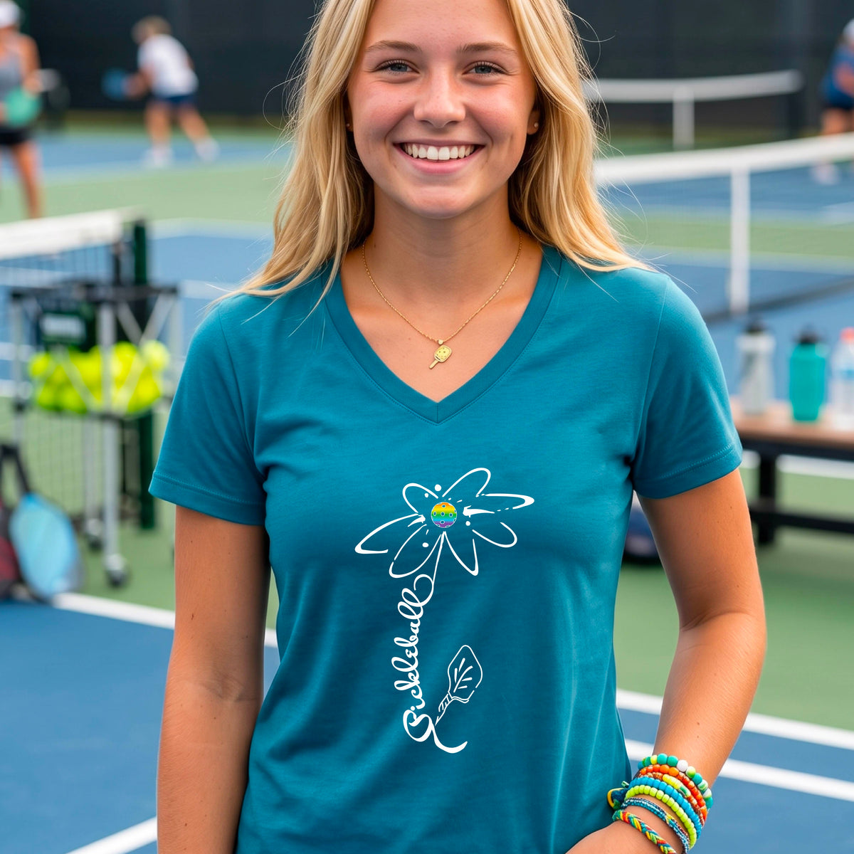 A woman wearing a teal short-sleeve V-Neck t-shirt with a pickleball graphic of a flower with a flower that has a colorful pickleball as the center and the stem is made of the word ‘PICKLEBALL’ and the leaf is a pickleball paddle, standing on a pickleball court.