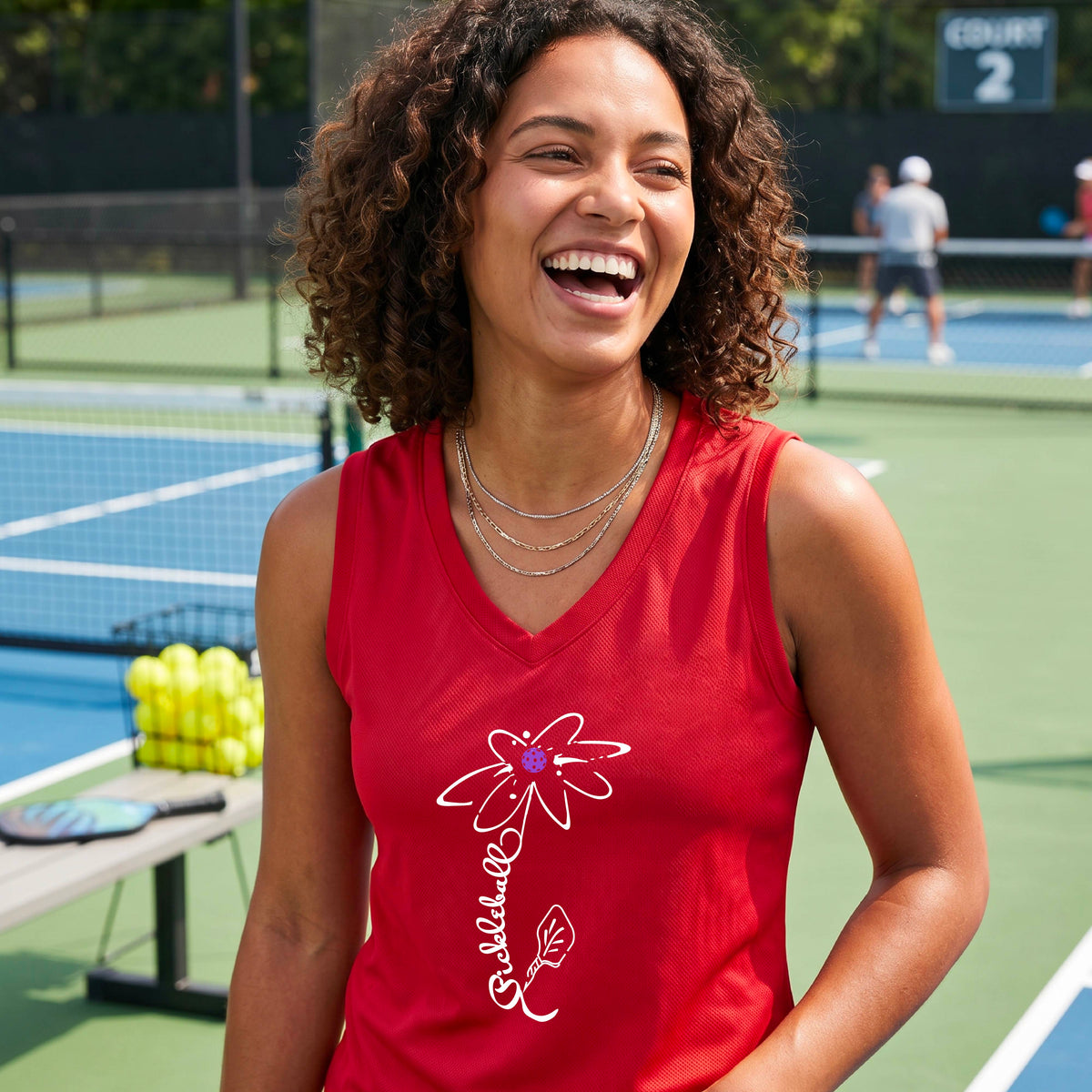 A smiling woman on a pickleball court wearing a red sleeveless V-neck athletic tank top with a white pickleball flower graphic with the stem made of th wrod 'PICKLEBALL' and the leaf is a pickleball paddle. 
