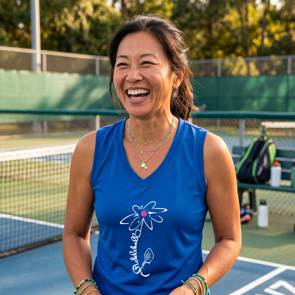 A smiling woman on a pickleball court wearing a royal blue sleeveless V-neck athletic tank top with a white pickleball flower graphic with the stem made of th wrod 'PICKLEBALL' and the leaf is a pickleball paddle. 
