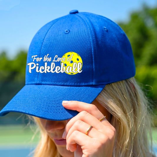 A close-up of a royal blue pickleball hat with a pickleball graphic of ‘FOR THE LOVE OF PICKLEBALL’ in white with a yellow pickleball behind it graphic on the front.
