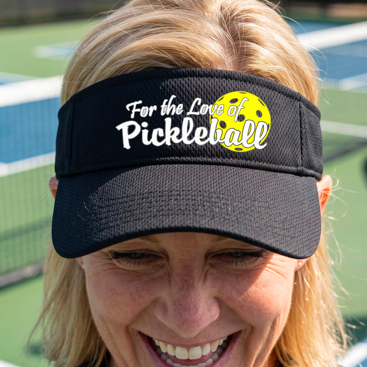 A close-up of a black pickleball visor with a pickleball graphic of ‘FOR THE LOVE OF PICKLEBALL’ in white with a yellow pickleball behind it graphic on the front.