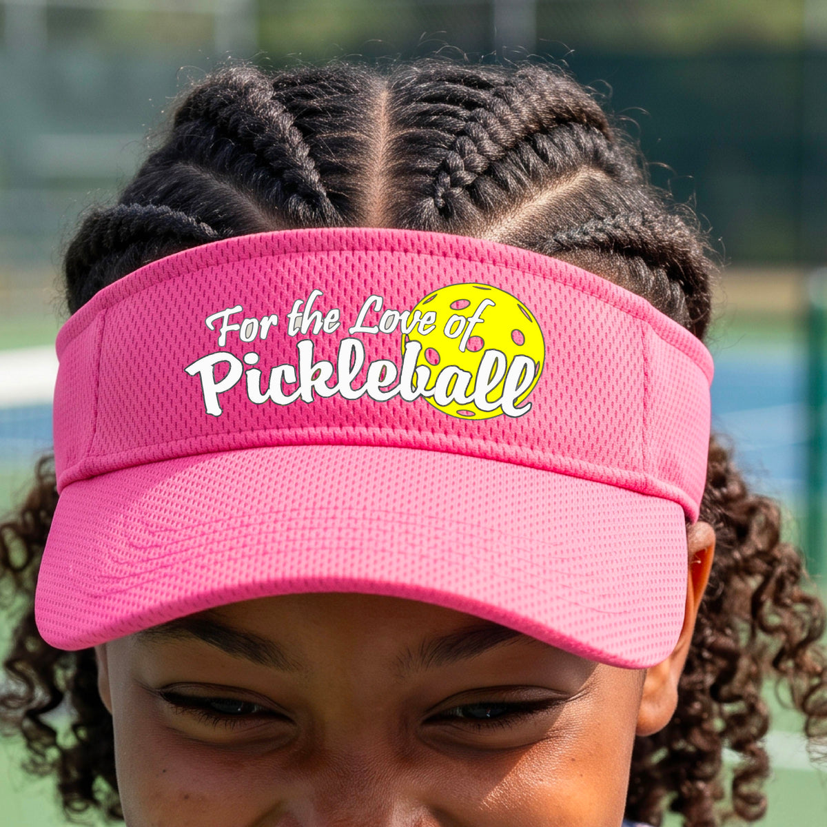 A close-up of a pink pickleball visor with a pickleball graphic of ‘FOR THE LOVE OF PICKLEBALL’ in white with a yellow pickleball behind it graphic on the front.