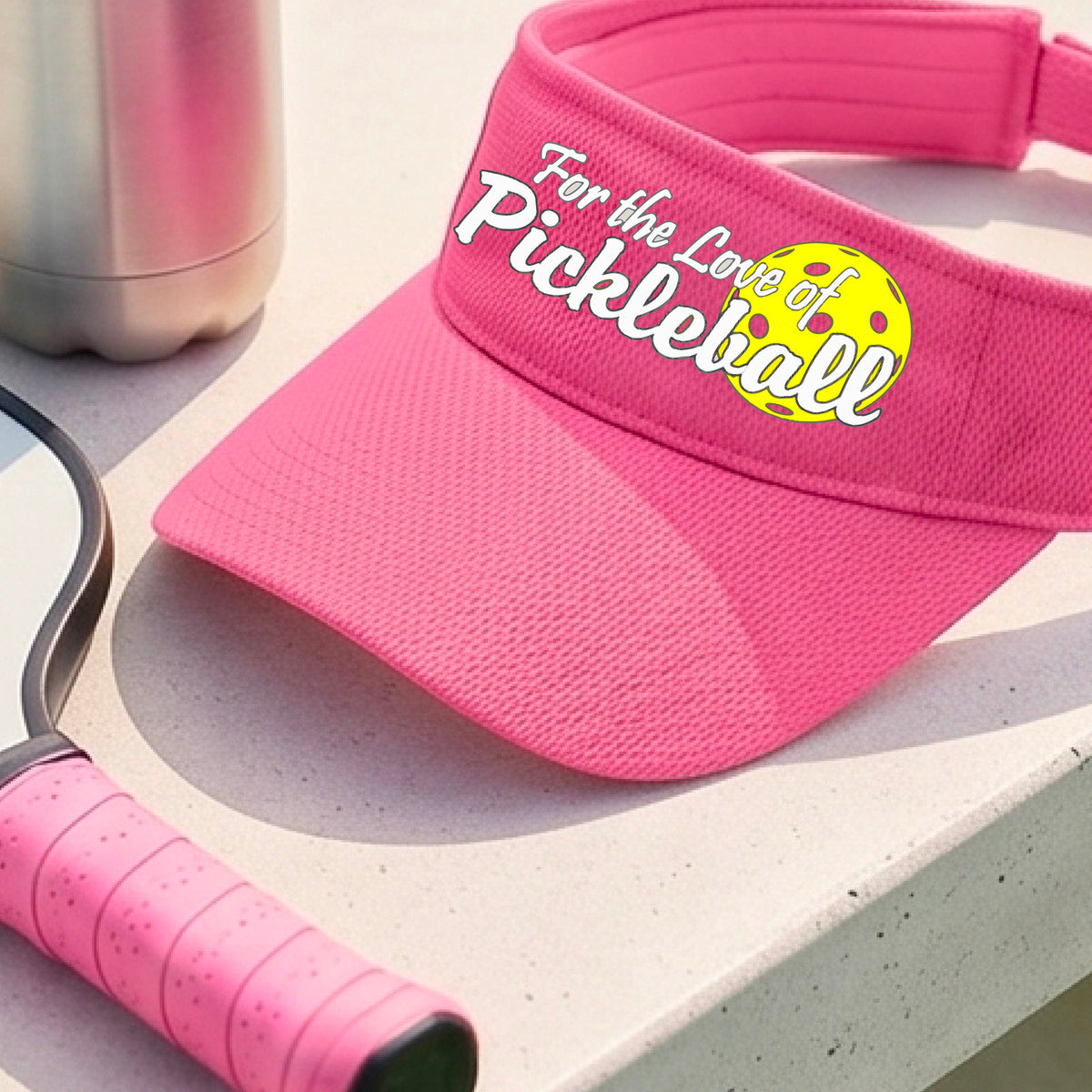 A close-up of a pink pickleball visor with a pickleball graphic of ‘FOR THE LOVE OF PICKLEBALL’ in white with a yellow pickleball behind it graphic on the front.