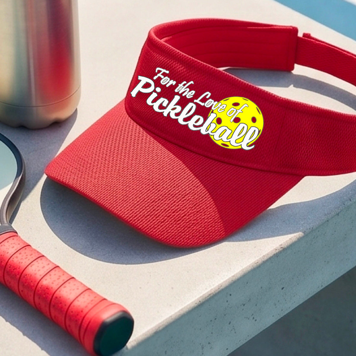 A close-up of a red pickleball visor with a pickleball graphic of ‘FOR THE LOVE OF PICKLEBALL’ in white with a yellow pickleball behind it graphic on the front.