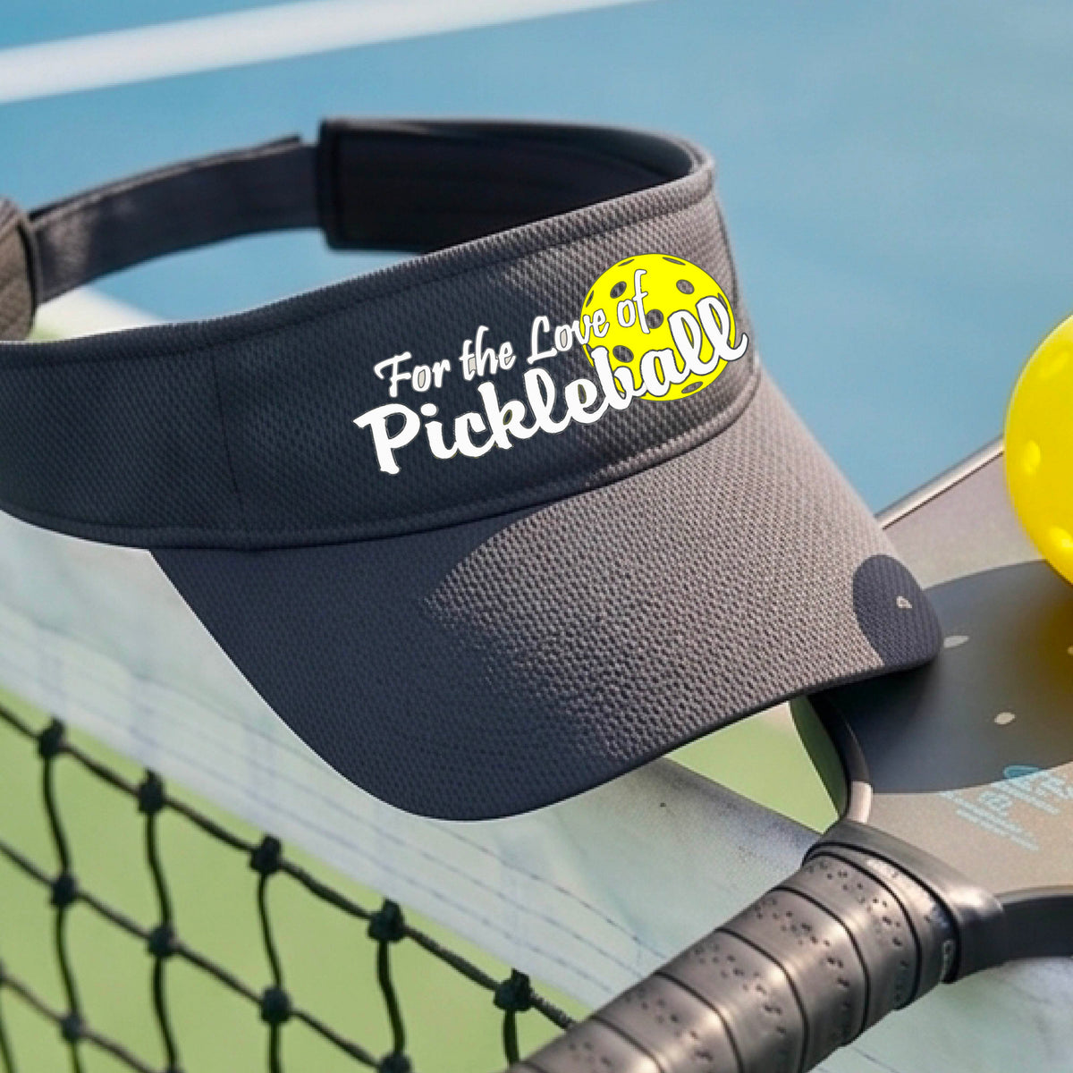 A close-up of a gray pickleball visor with a pickleball graphic of ‘FOR THE LOVE OF PICKLEBALL’ in white with a yellow pickleball behind it graphic on the front.