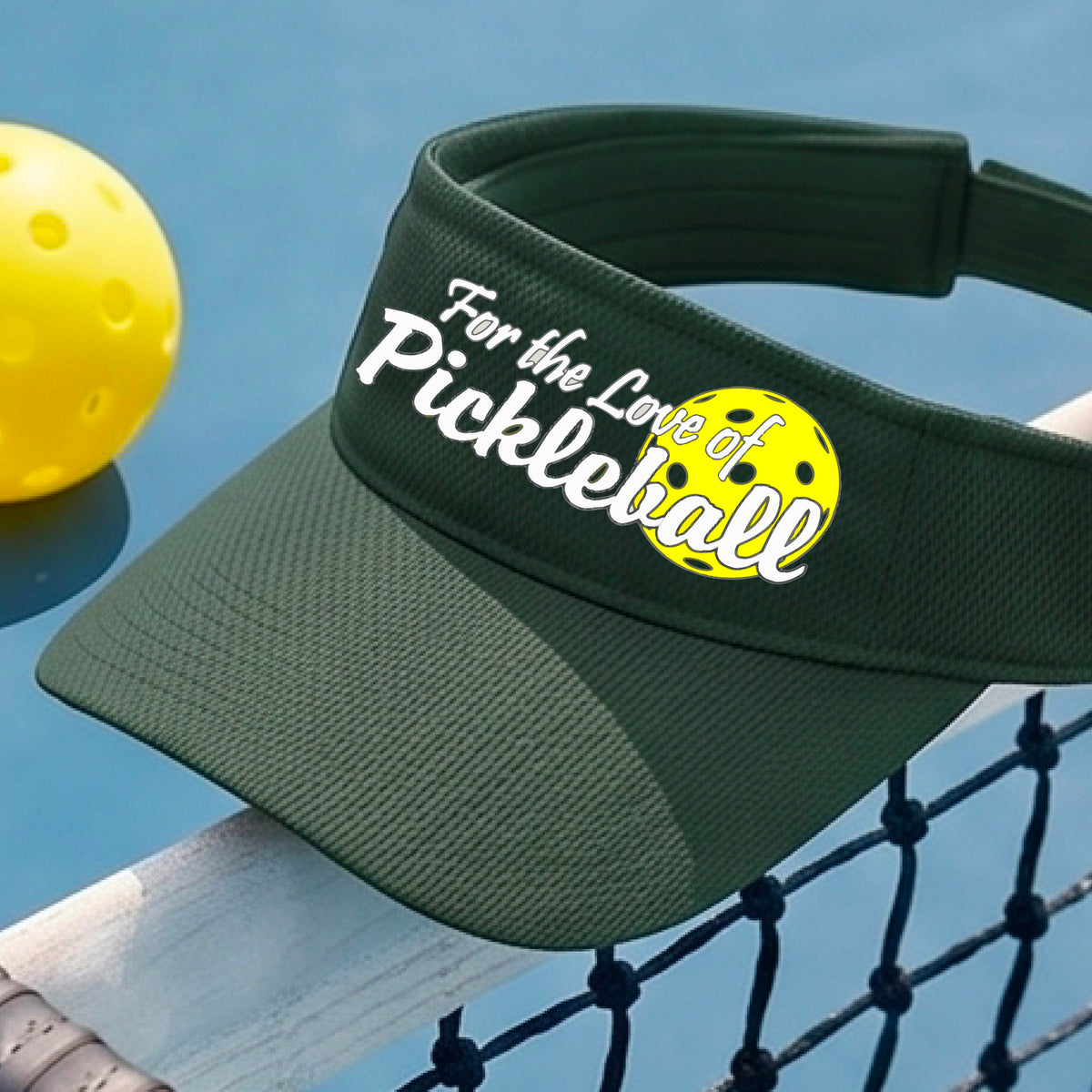 A close-up of a green pickleball visor with a pickleball graphic of ‘FOR THE LOVE OF PICKLEBALL’ in white with a yellow pickleball behind it graphic on the front.