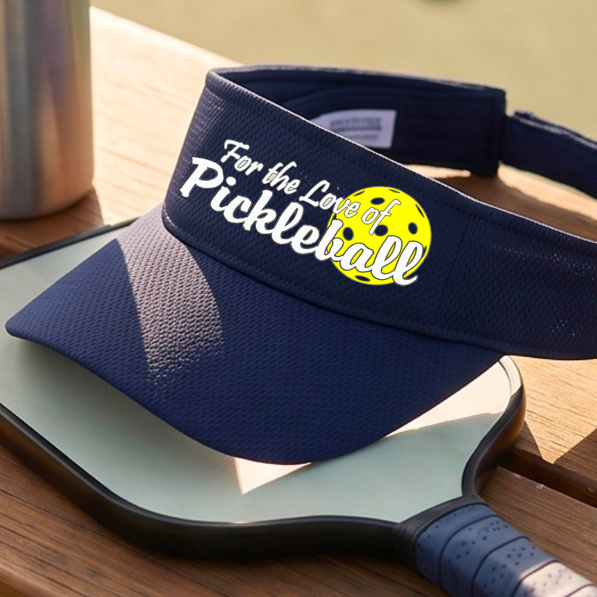 A close-up of a navy blue pickleball visor with a pickleball graphic of ‘FOR THE LOVE OF PICKLEBALL’ in white with a yellow pickleball behind it graphic on the front.
