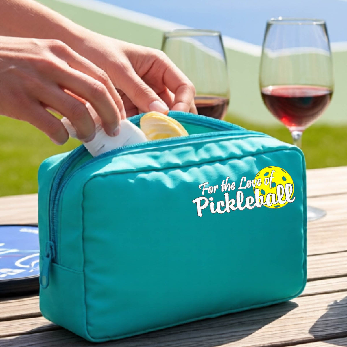 A close-up of a turquoise cosmetic toiletry bag with a pickleball graphic of ‘FOR THE LOVE OF PICKLEBALL’ in white with a yellow pickleball behind it.  