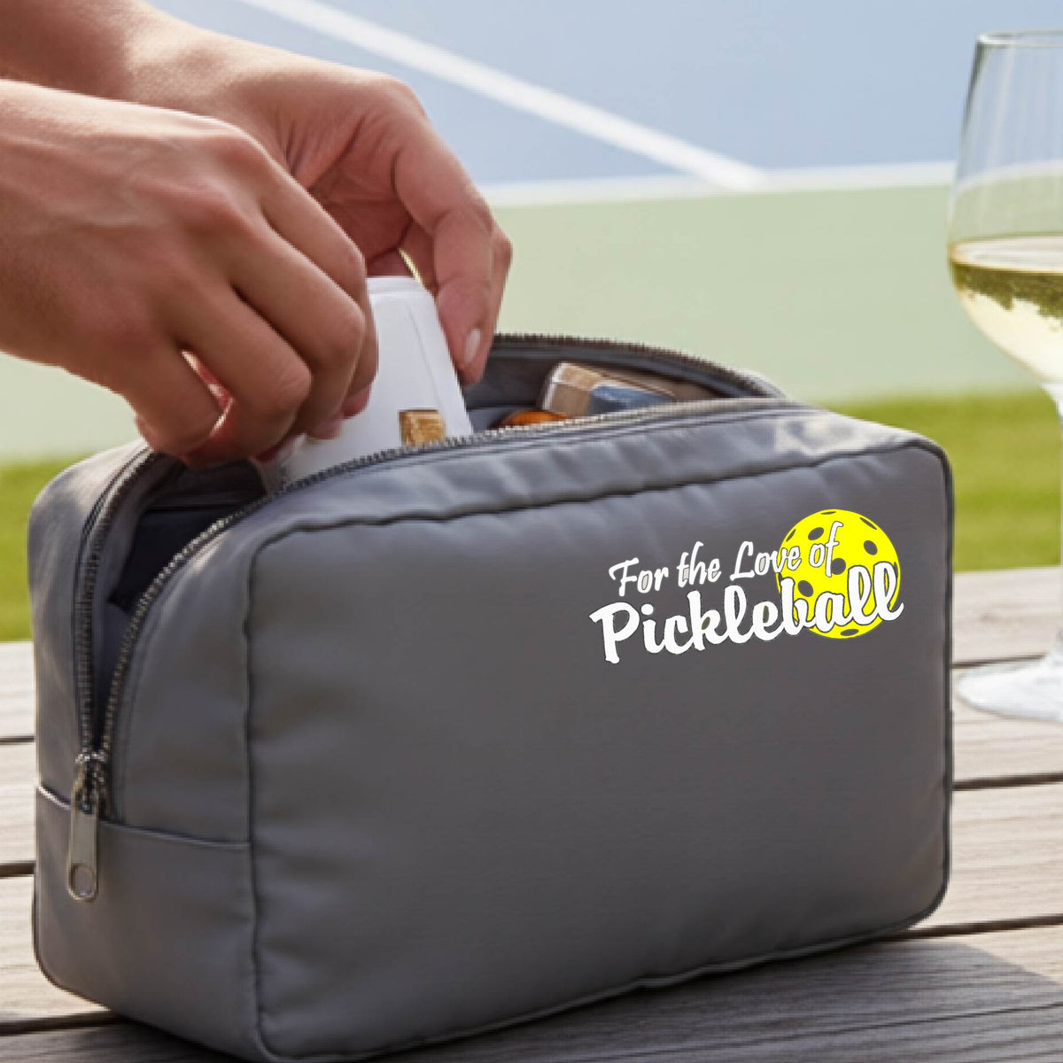 A close-up of a gray cosmetic toiletry bag with a pickleball graphic of ‘FOR THE LOVE OF PICKLEBALL’ in white with a yellow pickleball behind it.  
