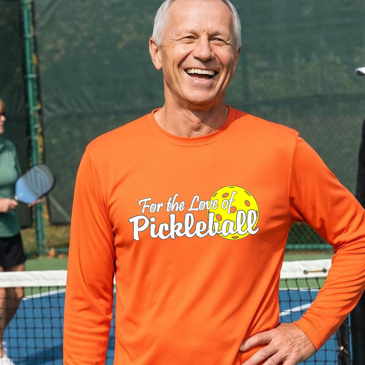 A close-up of a man wearing an orange long-sleeve t-shirt with a pickleball graphic on the left bottom left of ‘FOR THE LOVE OF PICKLEBALL’ in white with a yellow pickleball behind it.   