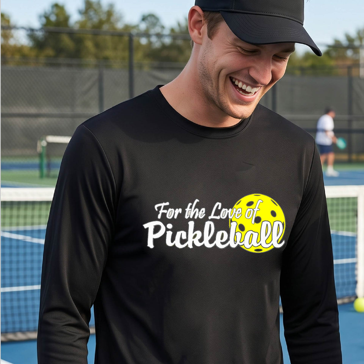 A close-up of a man wearing a black long-sleeve t-shirt with a pickleball graphic on the left bottom left of ‘FOR THE LOVE OF PICKLEBALL’ in white with a yellow pickleball behind it.  